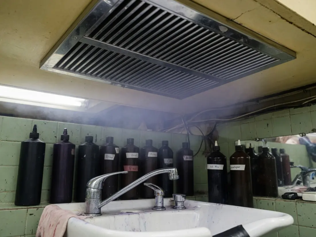 染发工位上方的金属排风口，水槽边一排深色药剂瓶，空气中有薄雾