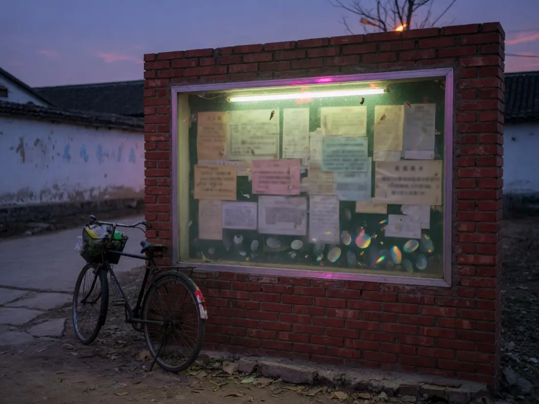 黄昏砖砌村务公示栏，灯管亮着，玻璃上有手印，自行车靠在旁边